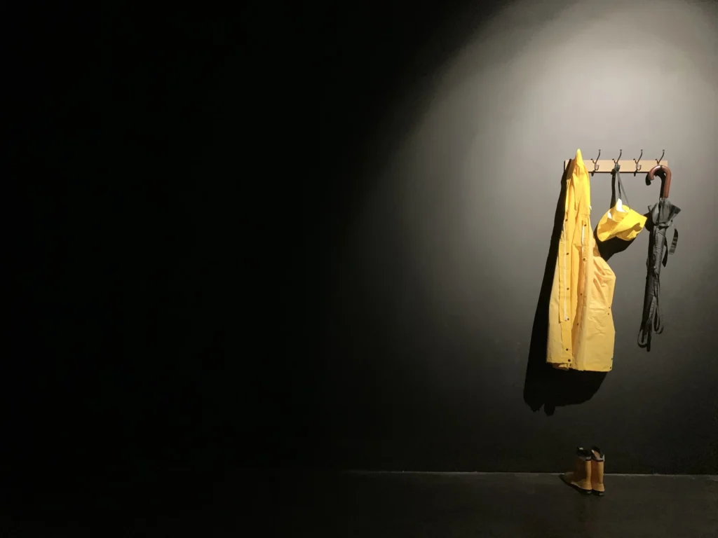 A yelloe souwester oon a coat hook on a black wall with yellow wellingtons on tje floor and an umbrella