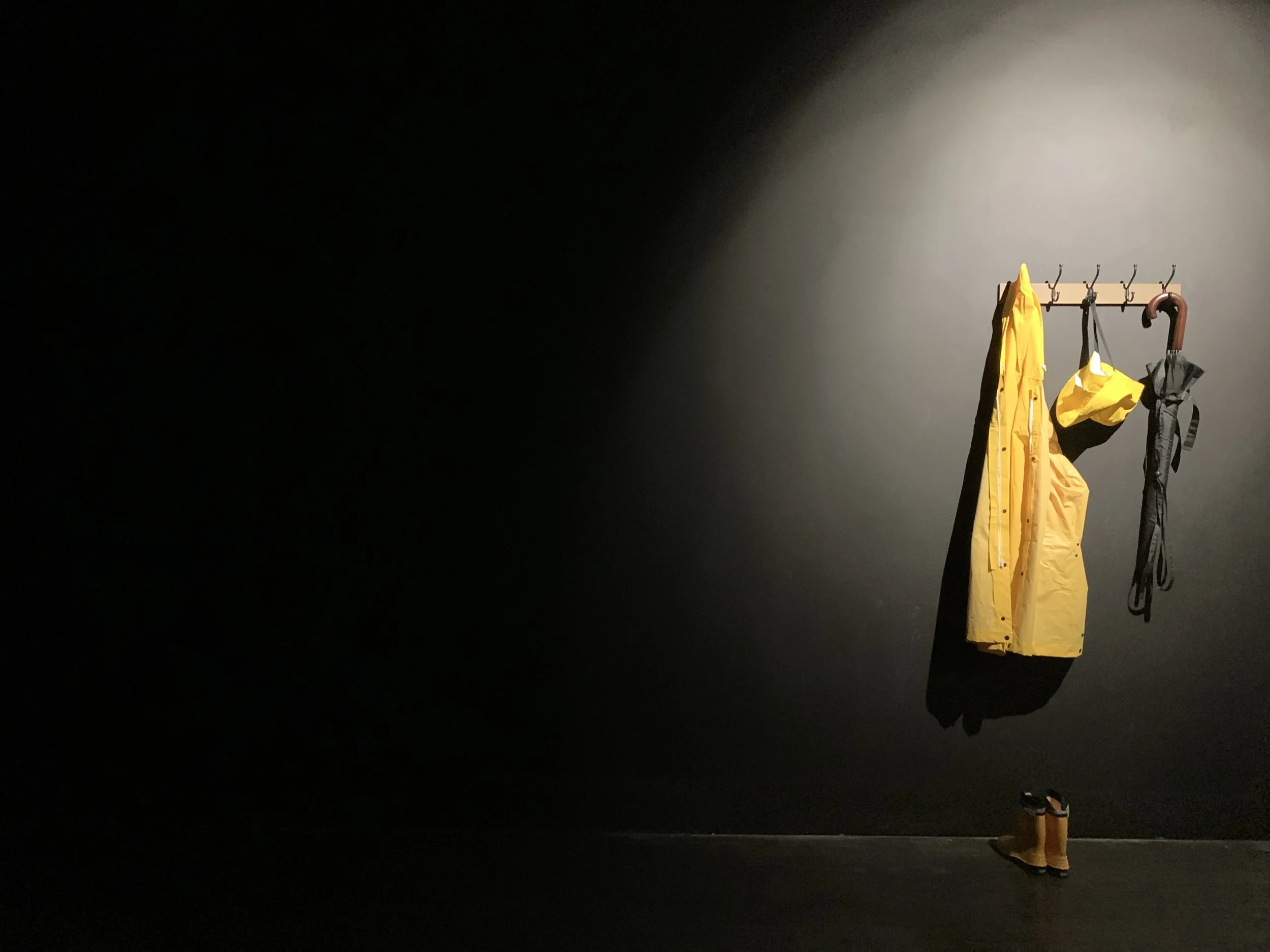 A yelloe souwester oon a coat hook on a black wall with yellow wellingtons on tje floor and an umbrella